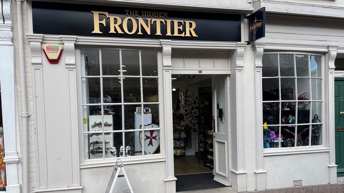 The storefront of 'The Hidden Frontier' shop, featuring a black sign with gold lettering, white brick paving, and a white shop facade with large multi-pane windows.